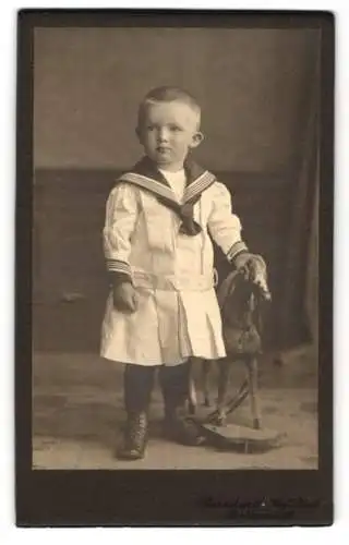 Fotografie Atelier Bernhard, Ballenstedt, Junger Knabe im Matrosenkleid mit Spielzeugpferd