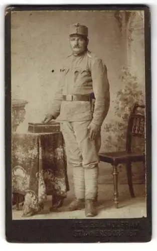 Fotografie A. Wolfram, Stammersdorf, Soldat in Uniform neben Tisch und Stuhl