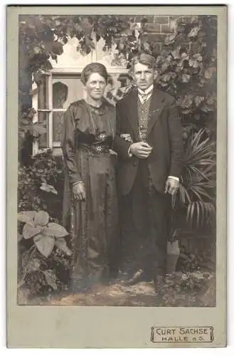 Fotografie Curt Sachse, Halle a. S., Ein elegantes Paar vor einem Fenster mit üppigem Blattwerk