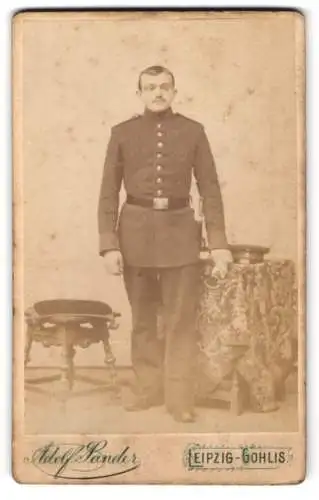 Fotografie Adolf Sander, Leipzig-Gohlis, Leipziger Strasse 12, Stehender Soldat in Uniform neben Tisch und Stuhl