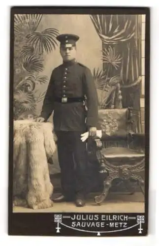 Fotografie Julius Eilrich, Metz, Ein Soldat in Uniform neben einem dekorativen Stuhl und einem Fell