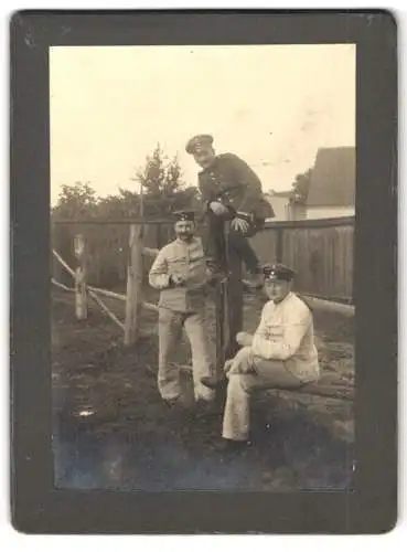 Fotografie unbekannter Fotograf und Ort, Uffz. in Uniform mit Sporen sitzt auf einem Zaun nebst zwei Soldaten