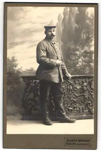 Fotografie Joseph Werner, München, Soldat in Feldgrau Uniform mit Krätzchen vor einer Studiokulisse