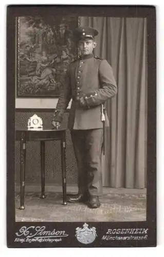 Fotografie X. Simson, Rosenheim, Münchenerstrasse 6, Soldat in Uniform neben Tisch mit Uhr