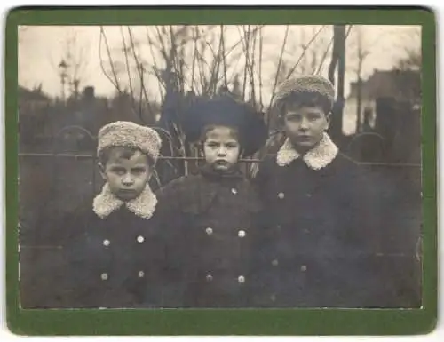 Fotografie unbekannter Fotograf und Ort, Drei Kinder in Winterkleidung vor einem Gartenzaun