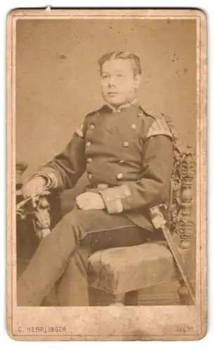 Fotografie C. Herrlinger, Ulm, beim Bahnhof, Uffz. Musiker in Uniform mit Bajonett & Schwalbennestern auf Stuhl sitzend