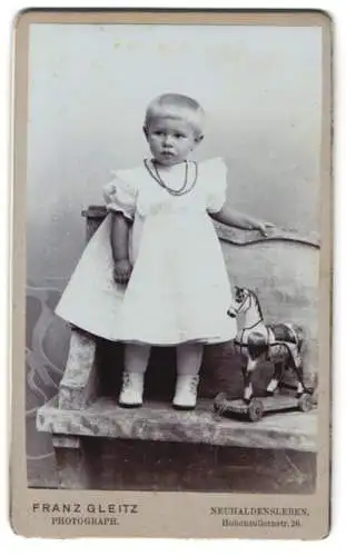 Fotografie Franz Gleitz, Neuhaldensleben, kleines blondes Mädchen im weissen Kleid mit Spielzeugpferd