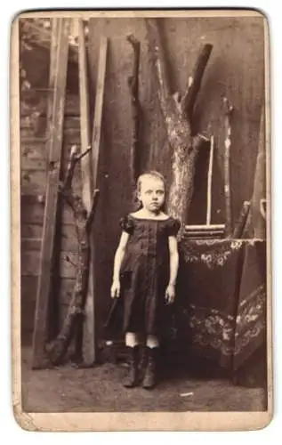 Fotografie unbekannter Fotograf und Ort, kleides Mädchen im dunklen Kleid vor einer Holzwerkstatt, Studiokulisse