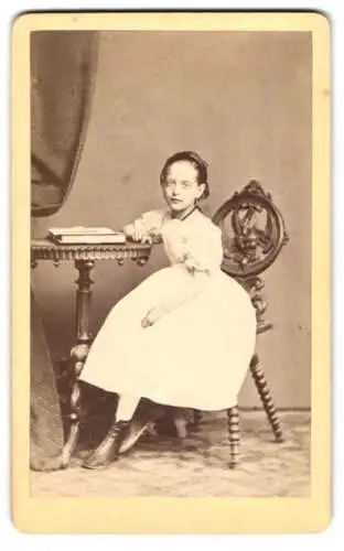 Fotografie unbekannter Fotograf und Ort, Portrait Mädchen im weissen Kleid blättert im Fotoalbbum