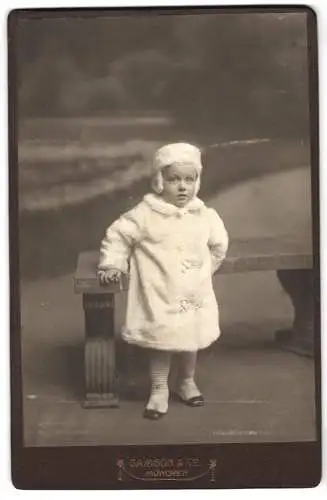 Fotografie Samson & Co., München, Neuhauserstr. 7, Kind in winterlicher Kleidung an einer Bank lehnend