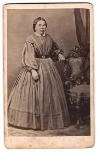Fotografie unbekannter Fotograf und Ort, Frau in elegantem Kleid neben kunstvollem Stuhl
