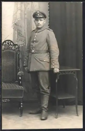 Foto-AK Soldat mit Schirmmütze in einer Stube, Uniformfoto