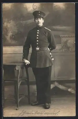 Foto-AK Soldat mit Schirmmütze, Quaste u. Handschuhen vor Fotokulisse, Uniformfoto