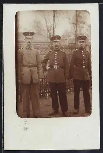 Foto-AK Drei Soldaten mit Schirmmützen vor einem Zaun, Uniformfoto