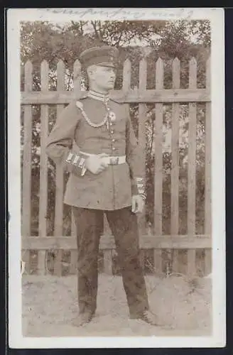 Foto-AK Soldat mit Schirmmütze, Kragenspiegel und Kette vor einem Lattenzaun, Uniformfoto