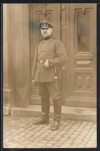 Foto-AK Soldat mit Schirmmütze und Quaste vor einem Gebäude, Uniformfoto