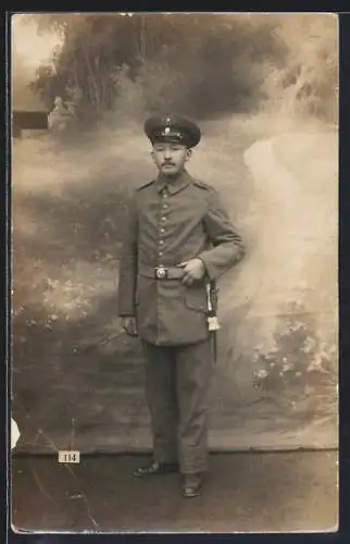 Foto-AK Soldat mit Schirmmütze und Quaste vor Fotokulisse, Uniformfoto
