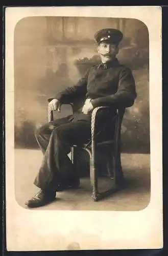 Foto-AK Soldat mit Schirmmütze im Sessel vor einer Fotokulisse, Uniformfoto