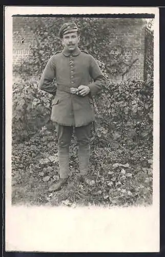 Foto-AK Soldat mit Schirmmütze u. Zigarre in einem Garten, Uniformfoto