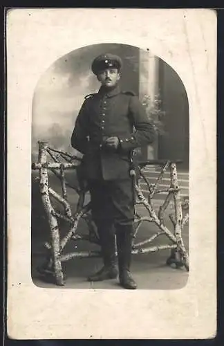 Foto-AK Soldat mit Schirmmütze u. Quaste vor Fotokulisse, Uniformfoto