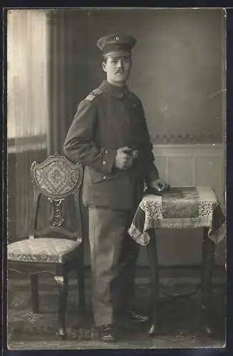 Foto-AK Soldat mit Schirmmütze in einer Stube, Uniformfoto