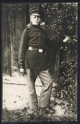 Foto-AK Soldat mit Schirmmütze in Zivilhosen vor einem Schuppen, Uniformfoto