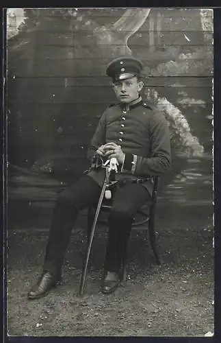Foto-AK Soldat mit Schirmmütze, Handschuhen u. Degen vor einer Holzwand, Uniformfoto