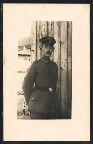 Foto-AK Soldat mit Schirmmütze und Brille vor einer Holzwand, Uniformfoto