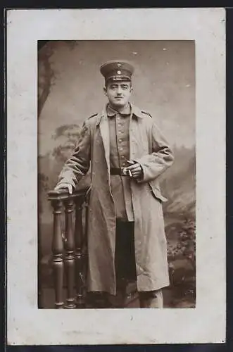 Foto-AK Soldat mit Schirmmütze im Mantel vor einer Fotokulisse, Uniformfoto