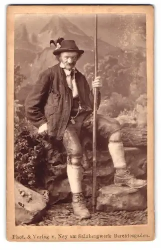 Fotografie Ney, Berchtesgaden, älterer Herr in bayerischer Tracht mit Lederhose und Wanderstab vor einer Studiokulisse