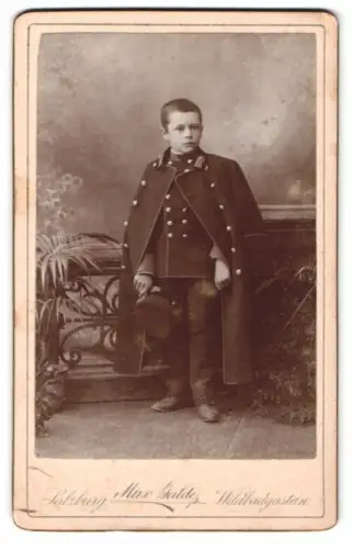 Fotografie Max Balde, Salzburg, kleiner junger Knabe als Kadett in Uniform mit Schirmmütze und Mantel