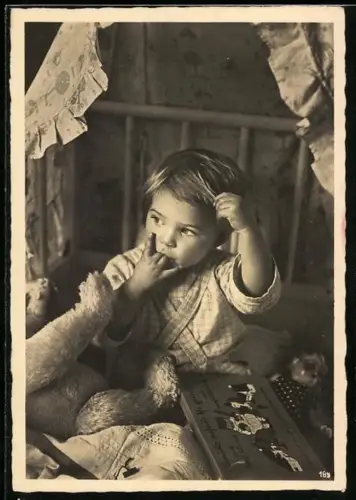 Foto-AK Kind sitzt mit Teddy und weiteren Spielzeugen in der Krippe