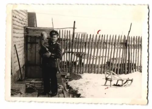 Fotografie junger NVA Soldat in Uniform mit Kind auf dem Arm, Schlitten im Schnee