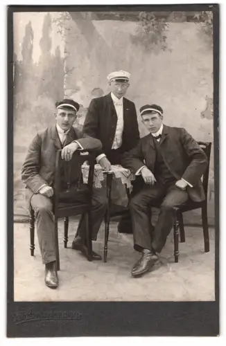 Fotografie Otto Brackmann, Obernkirchen, Drei Studenten in Anzügen und Mützen vor einer Studiokulisse, 1906