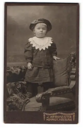 Fotografie J. Werkmeister, Freising, Ein Kind im eleganten Kleid auf einem dekorativen Sessel