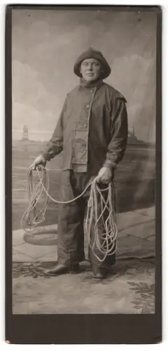 Fotografie unbekannter Fotograf und Ort, Fischer in Ölzeug mit Seil in der Hand vor einer Studiokulisse
