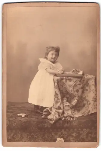 Fotografie unbekannter Fotograf und Ort, Kleines Kind im weissen Kleid an Tisch mit Blumen