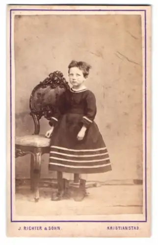 Fotografie J. Richter & Sohn, Kristianstad, kleines Mädchen dunklen Kleid neben verziertem Stuhl