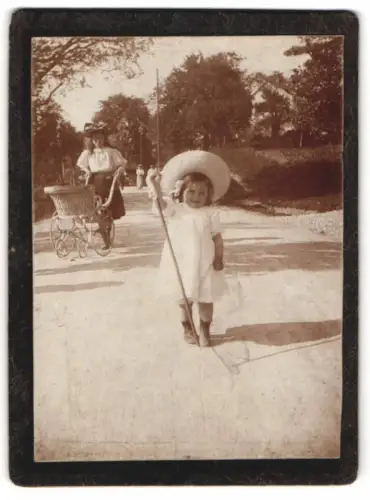 Fotografie unbekannter Fotograf und Ort, Kind mit grossem Hut auf Spaziergang, grosse Schwester mit Kinderwagen