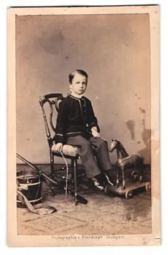 Fotografie Brandseph, Stuttgart, junger Knabe im Anzug mit Holzspielzeug Pferd und Gewehr samt Trommel