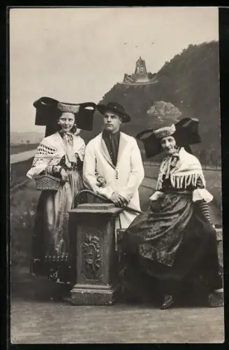 Foto-AK Herr mit zwei Frauen in Tracht, Im Hintergrund Denkmal, Studiokulisse