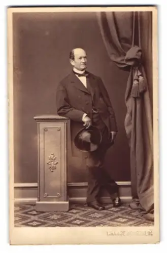 Fotografie L. Haase & Co., Berlin, Herr im Anzug mit Fliege und Zylinder lehnt an einem Podest