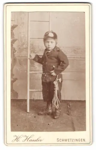 Fotografie H. Hassler, Schwetzingen, niedlicher junger Knabe als Feuerwehrmann in Uniform mit Helm und Leiter
