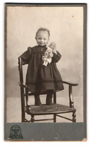 Fotografie Hermann Tietz, Berlin, niedliches kleines Mädchen stehend auf dem Stuhl mit Puppe im Arm