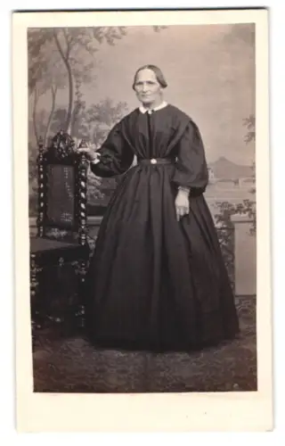 Fotografie unbekannter Fotograf und Ort, Frau in dunklem Kleid neben Stuhl