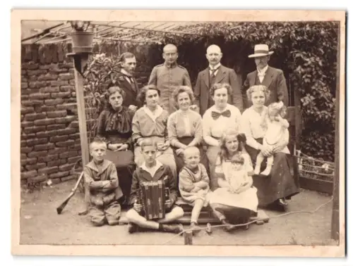 Fotografie unbekannter Fotograf und Ort, Gruppenportrait im Garten Knabe mit Akkordeon und Puppe