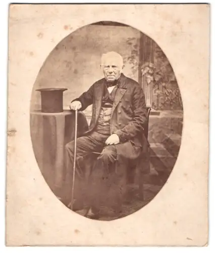 Fotografie unbekannter Fotograf und Ort, älterer Herr im Anzug mit Stock und Zylinder auf dem Tisch, 1861