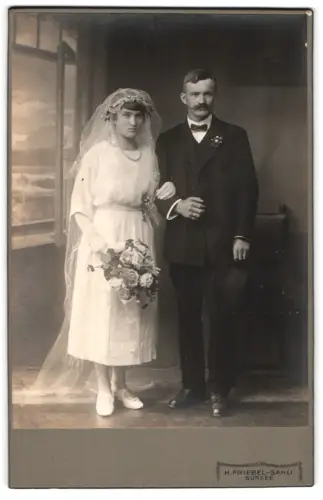 Fotografie H. Friebel-Sahli, Sursee, Brautpaar im Hochzeitskleid und Anzug vor einer Studiokulisse