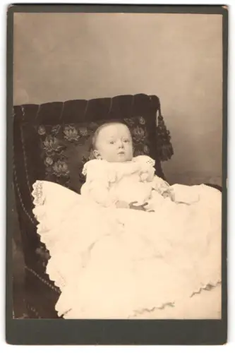 Fotografie unbekannter Fotograf und Ort, Baby im Taufkleid auf einem Sessel liegend