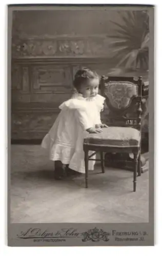 Fotografie A. Dilger & Sohn, Freiburg, Rheinstrasse 30, Kleinkind in weissem Kleid neben Stuhl